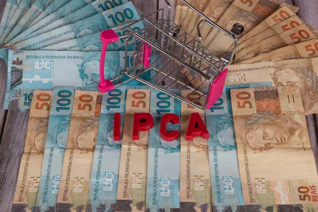 Cédulas de Real (50 e 100 reais) espalhadas sob a sigla IPCA em letras vermelhas e um pequeno carrinho de compras de metal tombado, ilustrando o impacto da inflação no poder de compra.