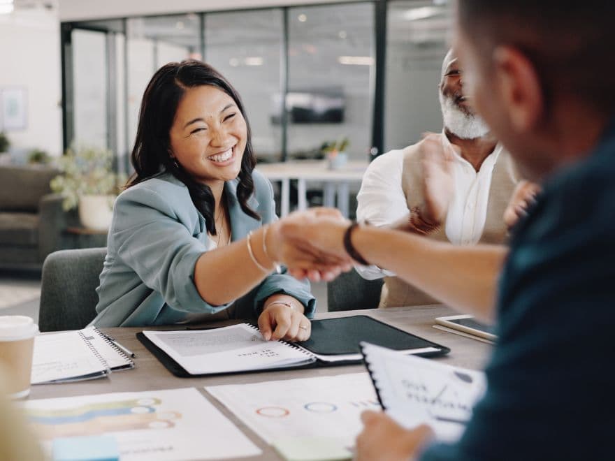 Uma corretora de imóveis sorrindo e apertando a mão de um cliente em uma mesa de reunião, simbolizando o fechamento de uma negociação imobiliária de sucesso.