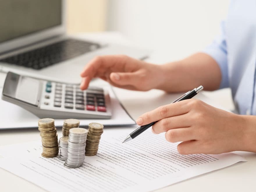 Corretor de imóveis usando calculadora sobre documentos com pilhas de moedas ao lado, representando o cálculo de outorga onerosa.