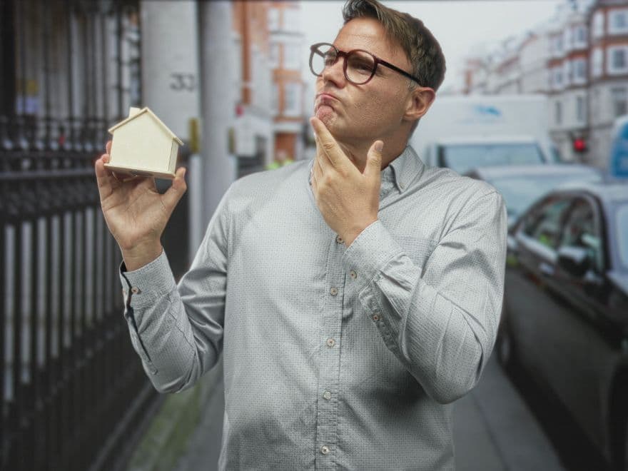 Homem na rua com expressão facial de dúvida, com a mão no queixo e segurando uma casinha decorativa.