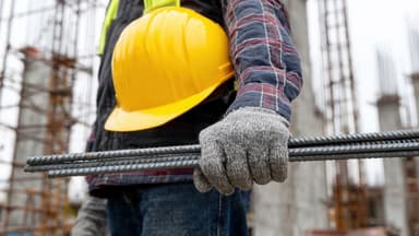 Trabalhador da construção civil segurando barras de vergalhão nervurado em um canteiro de obras.