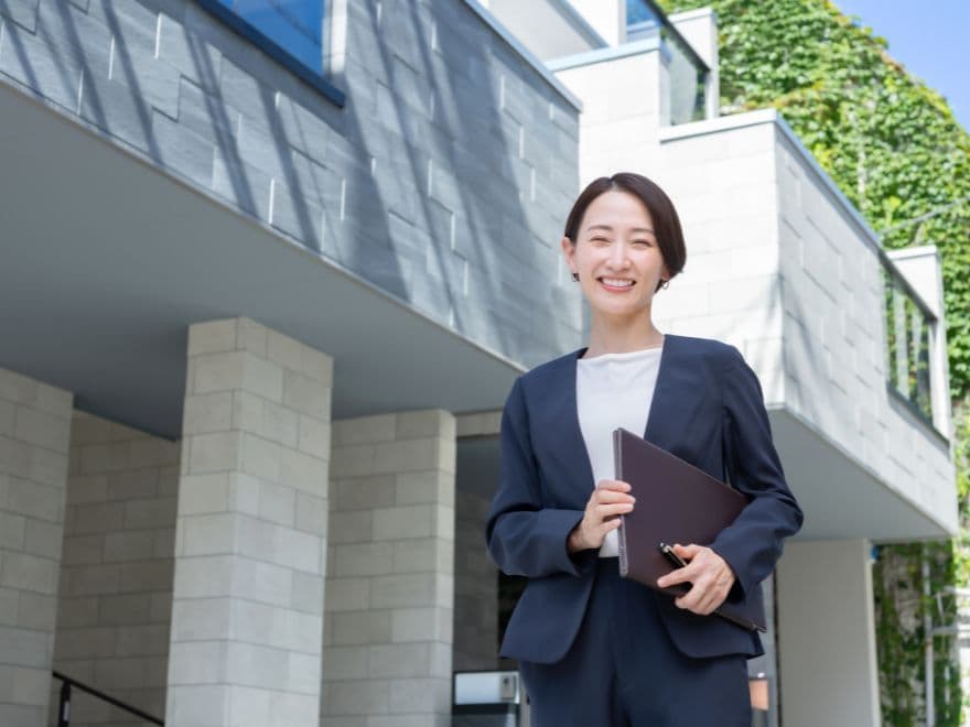 Uma corretora de imóveis sorridente, segurando uma pasta de documentos em frente a um edifício de alto padrão, conduzindo uma avaliação de imóveis.