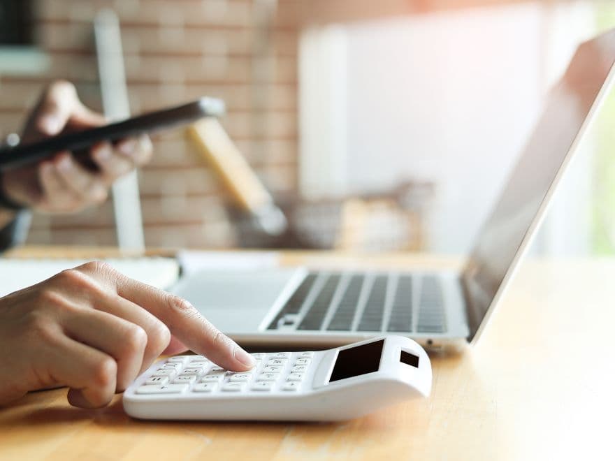 Pessoa mexendo no celular, enquanto há um notebook sob a mesa, junto com a calculadora que ela digita. Representando uma análise de faturamento e lucro.